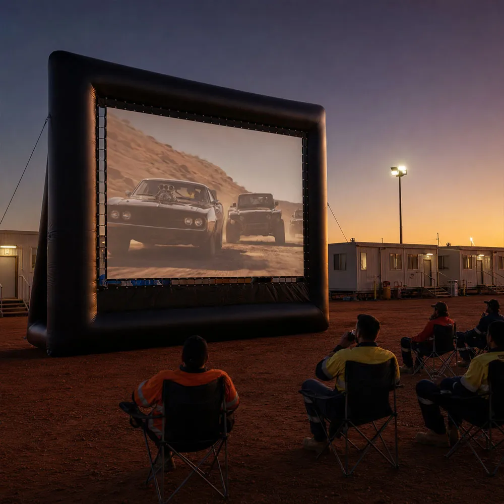 An inflatable outdoor cinema screen at a remote mining camp at dusk, hi-vis crew watching from camp chairs on red dirt