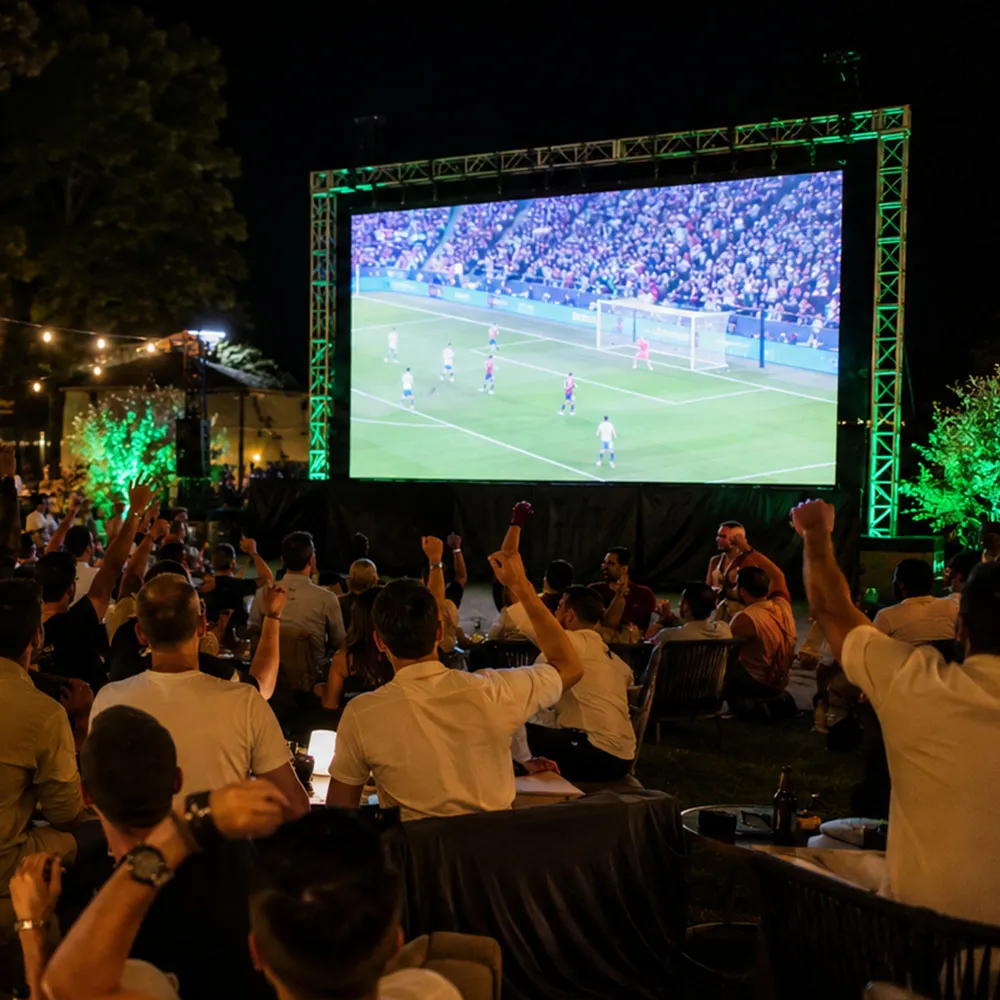 A crowd watching live sport on a big outdoor screen at night